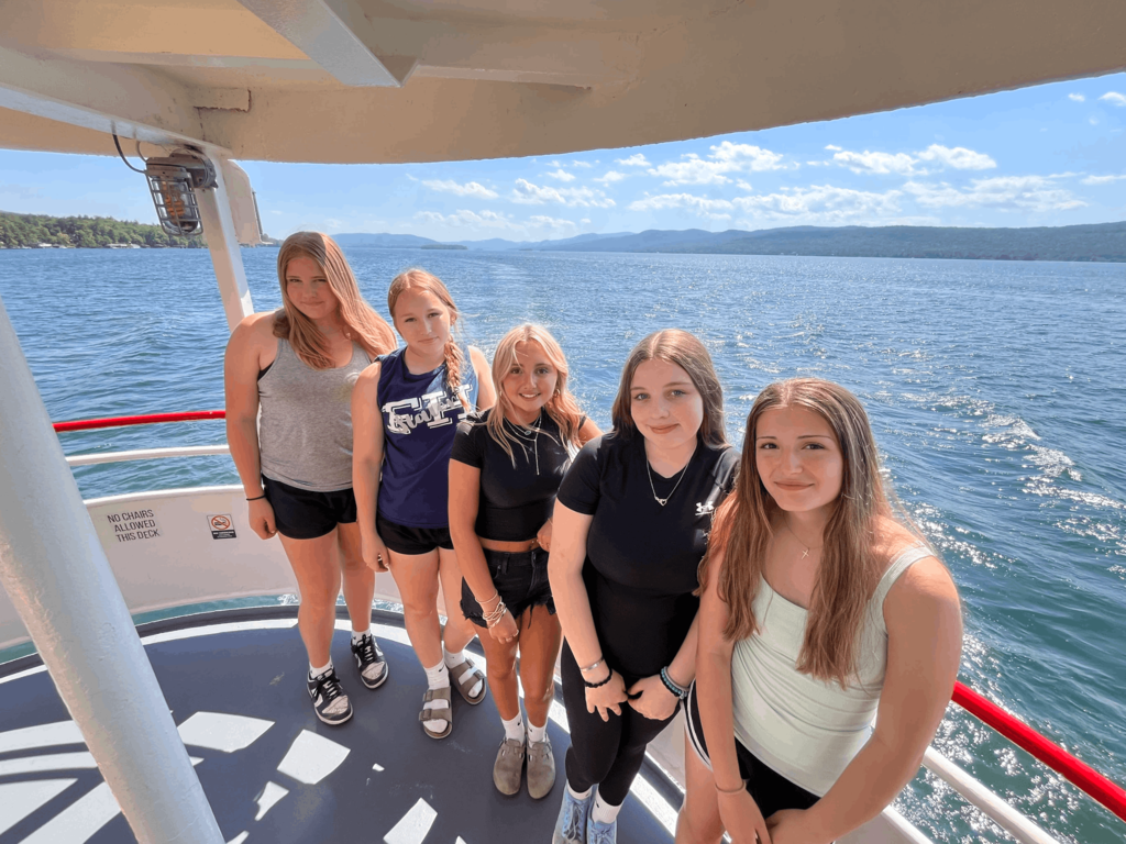 Five students pose together on the deck of a boat cruising on Lake George, with mountains and blue water in the background.