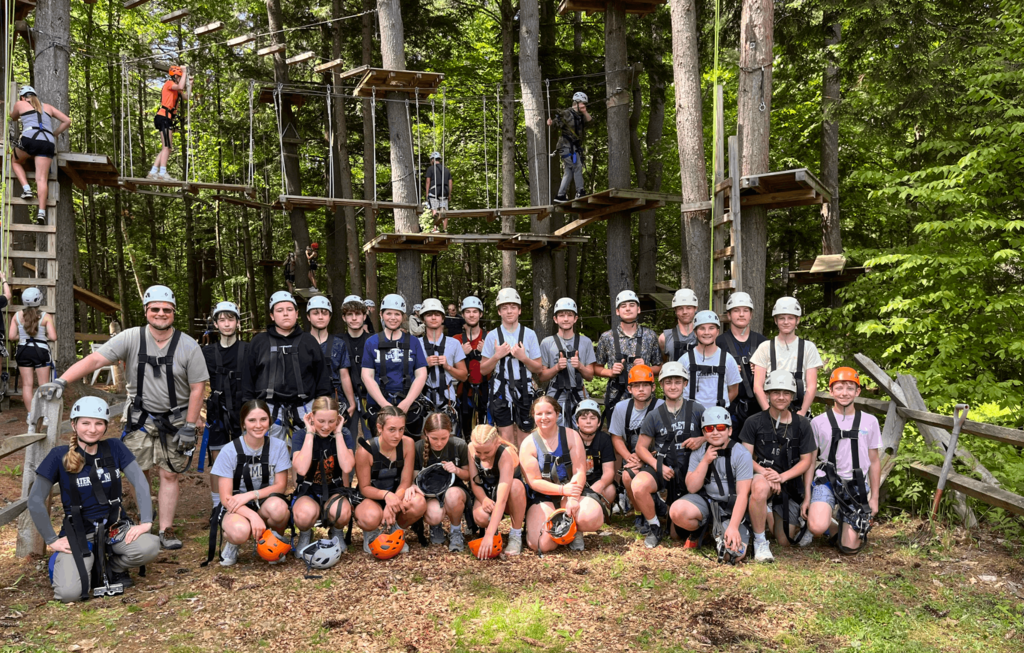 Group photo of students and staff wearing helmets and safety harnesses at an outdoor aerial ropes course, with participants climbing and crossing obstacles in the background.
