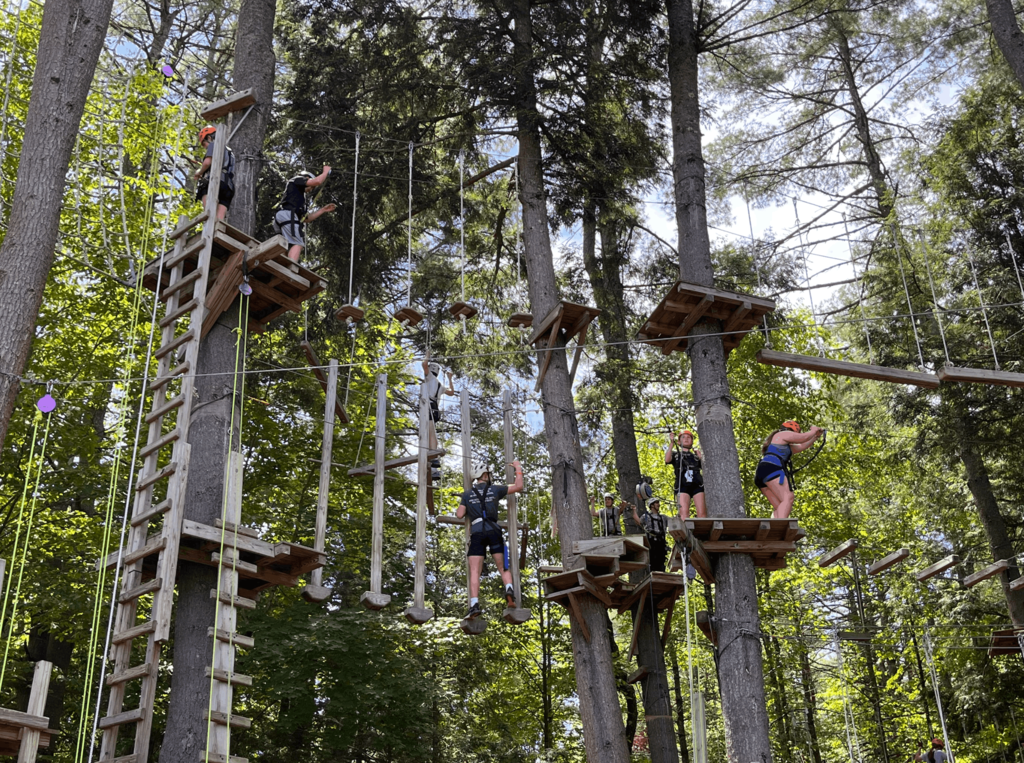 Students navigate multiple elevated ropes course obstacles and platforms among tall trees during an outdoor adventure activity.