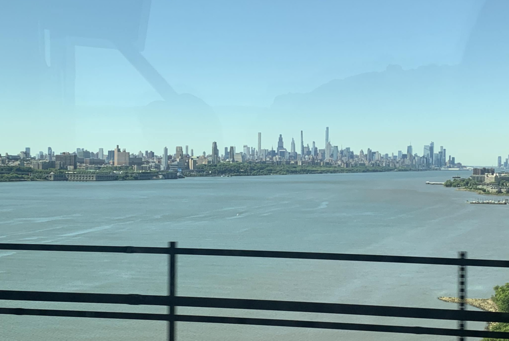 View of the Manhattan skyline across a wide river, photographed from a moving vehicle crossing a bridge. Skyscrapers rise in the distance against a clear blue sky.