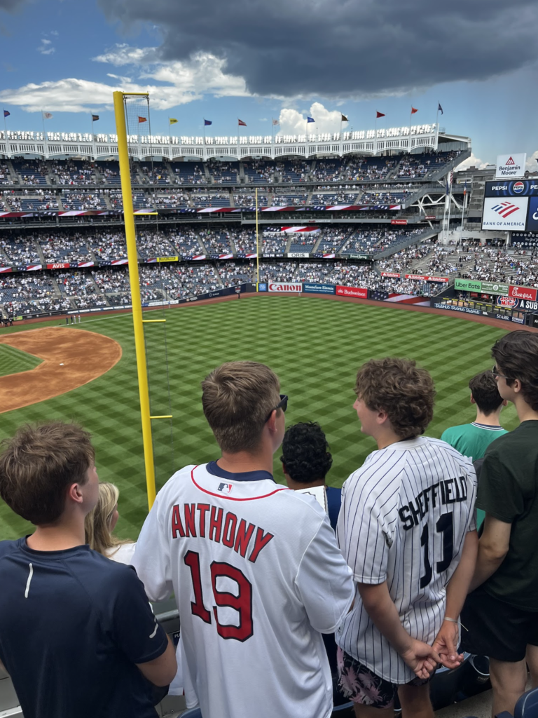 Students stand along the railing at Yankee Stadium watching the game from seats near the outfield. Two students wear baseball jerseys while dark clouds gather overhead.