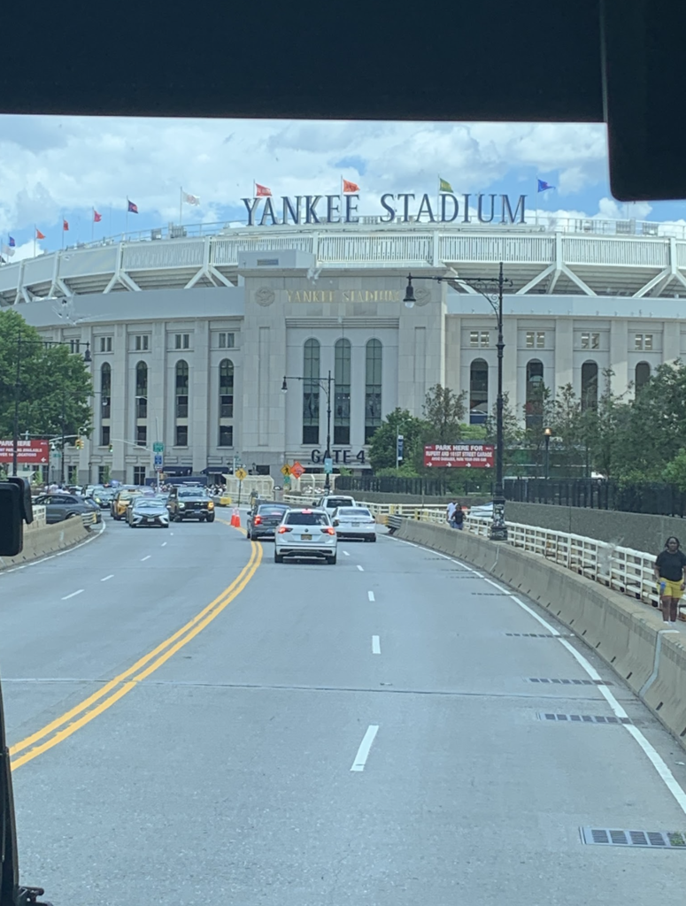 Exterior view of Yankee Stadium from a roadway approaching the stadium. Cars travel toward the entrance while the large "Yankee Stadium" sign is visible above the building.