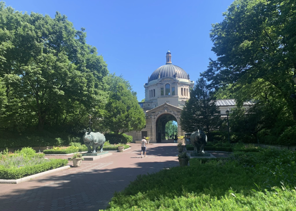 View of the entrance to the Bronx Zoo featuring a large domed historic building framed by trees and landscaped gardens on a sunny day.