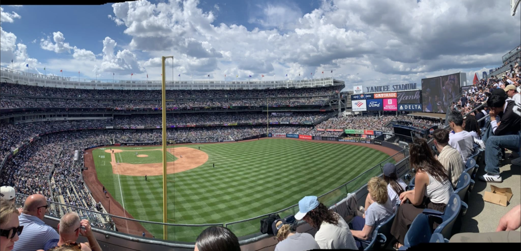 Wide panoramic view of Yankee Stadium during a baseball game. Thousands of fans fill the stands surrounding the field under a sky filled with large white clouds.