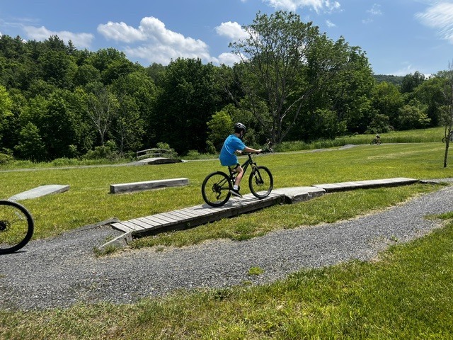 A student rides a mountain bike across a wooden balance feature on a skills course. The trail area includes gravel paths, grassy fields, and additional riding obstacles.