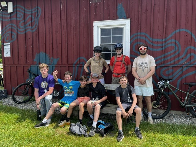 A group of students and adult leaders pose with their bicycles outside a red barn-style building on a sunny day. Several bikes are parked nearby as the group takes a break before or after a ride.