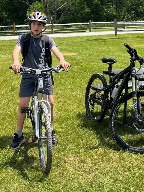 A student wearing a bicycle helmet and backpack sits on a mountain bike in a grassy field. Another bike is parked beside them on a sunny day.