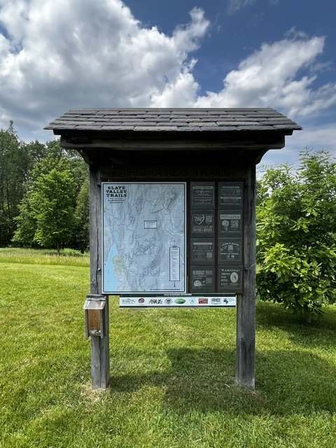 A trailhead information kiosk stands in a grassy field under a partly cloudy sky. The sign displays a map and information for the Slate Valley Trails Fairgrounds Trails network.