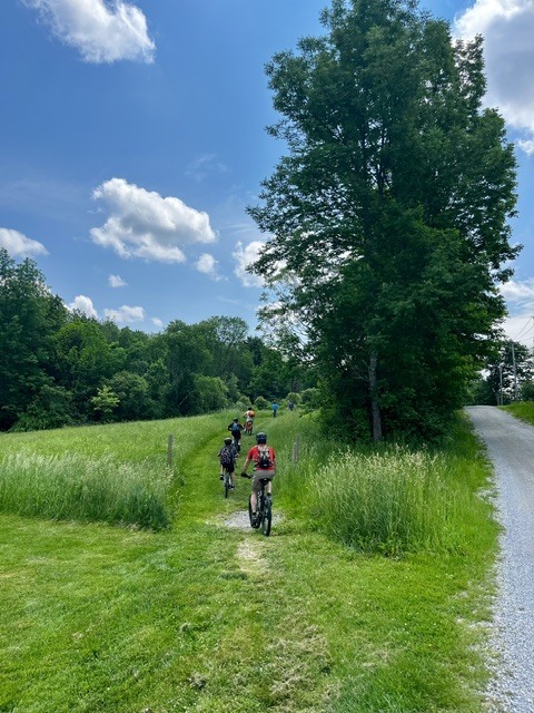 A line of student cyclists rides along a grassy trail bordered by tall grass and trees. The group heads toward a wooded area under a bright blue sky with scattered clouds.