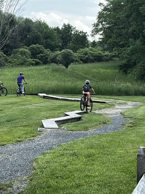 Two cyclists navigate a winding bike skills course featuring narrow wooden boardwalk sections and gravel paths. Trees and open grassy fields surround the riding area.
