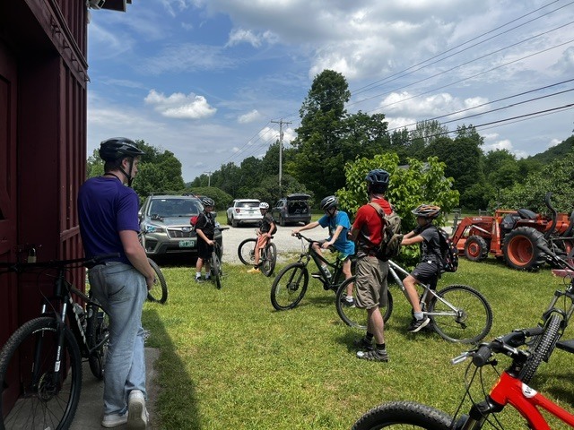 Students wearing helmets gather with their bicycles beside a red building and parking area. Adult leaders stand nearby as the group prepares for a trail ride.