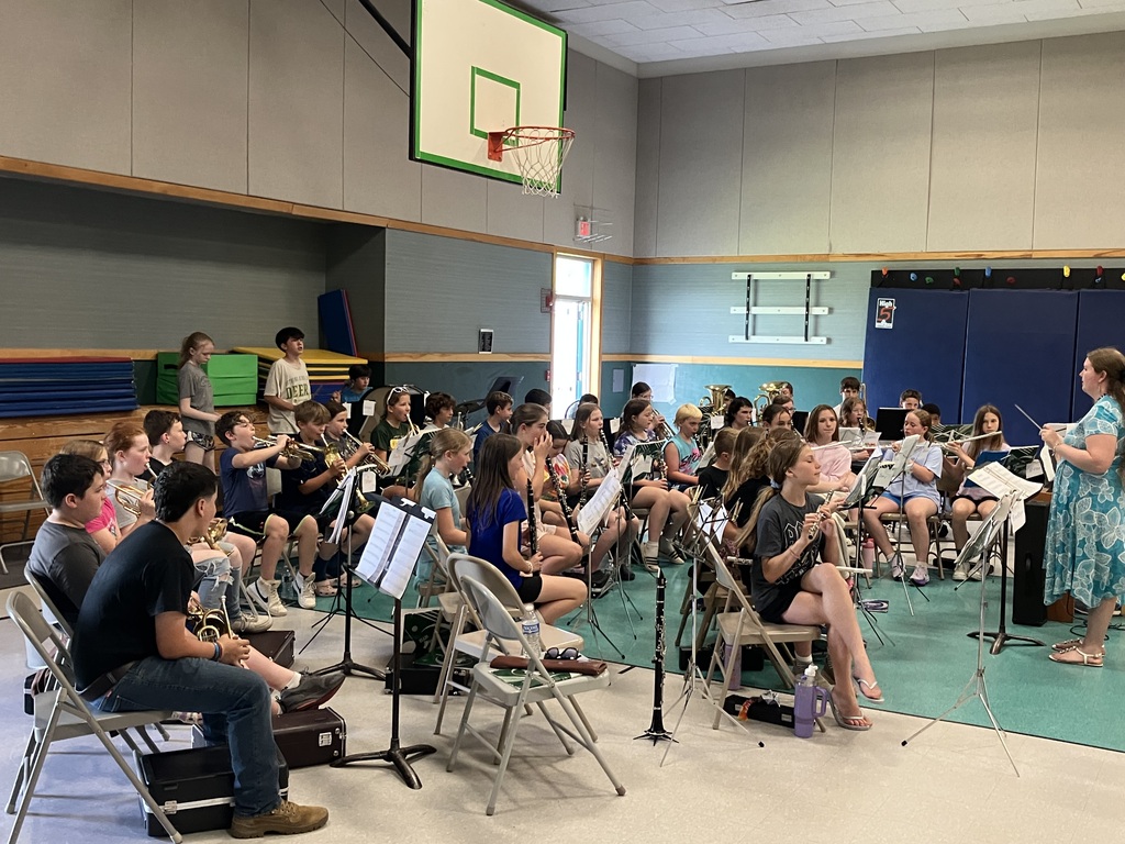 A close-up shot focusing on the brass section of a student band seated on folding chairs in a gymnasium. In the foreground, a boy wearing a black T-shirt, blue jeans, and tan boots holds a trumpet. Next to him, other students are visible holding trumpets and cornets, some chatting or adjusting their instruments. Black instrument cases are scattered on the floor beneath their chairs, and blue and green gym mats are stacked in the background. IMG_3421.jpg Alt Text: An angled shot of a student band rehearsing in a school gym under a basketball hoop. A conductor with long hair wearing a blue patterned dress stands on the right, raising a baton to direct the musicians. The band includes students playing flutes, clarinets, and trumpets, all seated on folding chairs with music stands in front of them. Instrument cases rest on the floor nearby.