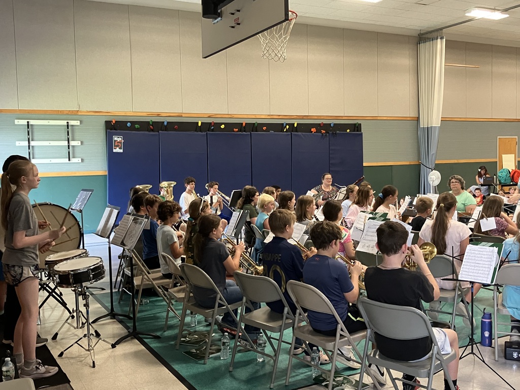 IMG_3418.jpg Alt Text: A wide, eye-level shot inside a school gymnasium where a student band is performing for an audience of younger children. The band students are seated in a large semi-circle on a teal mat under a basketball hoop, playing various woodwind and brass instruments. A female conductor in a green shirt and jeans stands at the center facing them. In the foreground, a large group of elementary school students are sitting cross-legged on the floor, watching the performance. Several adults are seated at cafeteria tables on the right side of the gym.
