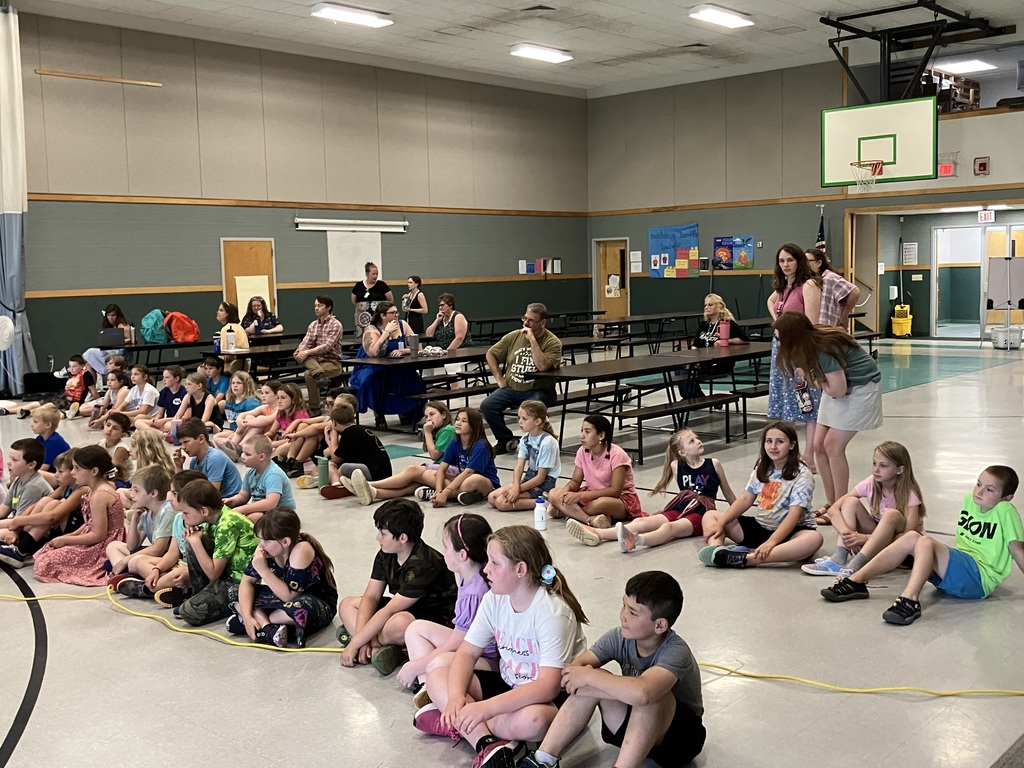 A wide shot focusing on the audience inside a school gymnasium turned cafeteria. Dozens of elementary-aged children are sitting cross-legged on the floor in rows, facing toward an unseen performance on the left. In the background, several adults and older students are seated at long brown cafeteria tables, watching the event. A scoreboard labeled "Benson Village School" is visible on the far wall.