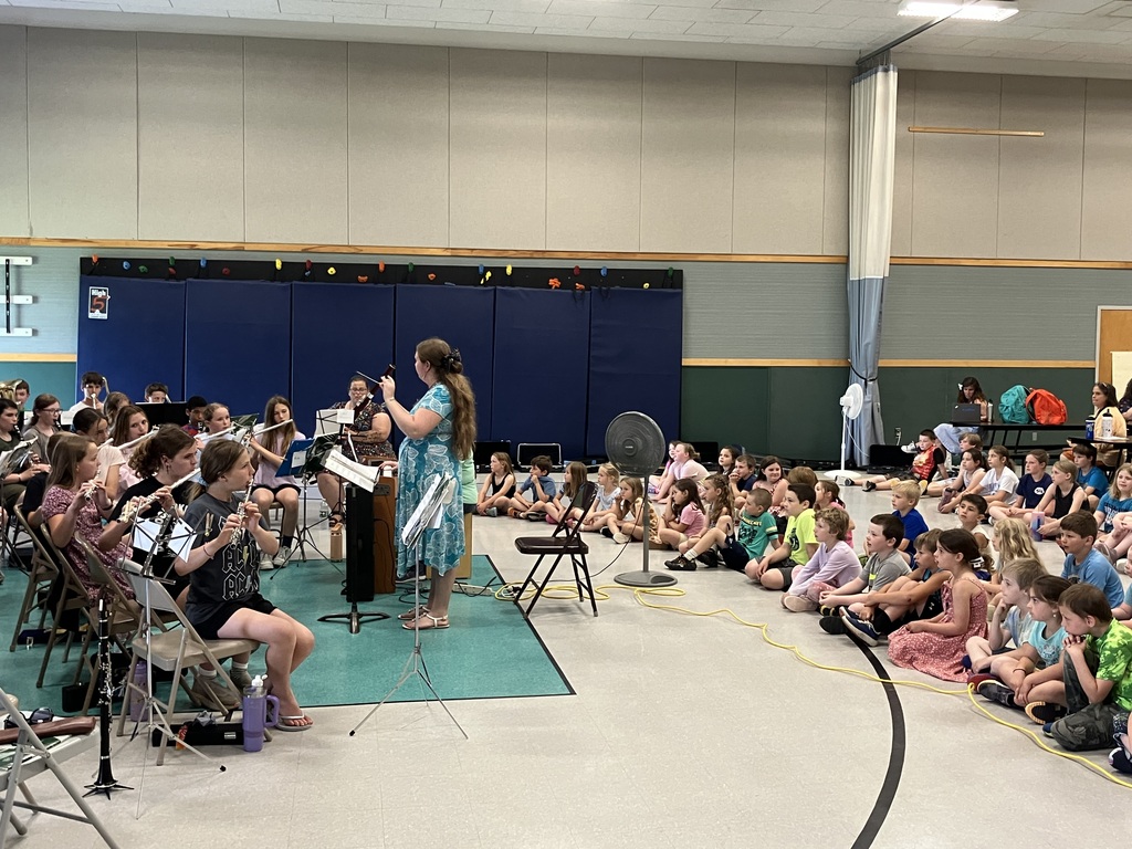 A high-angle, wide shot from the side of a school gymnasium during a band demonstration. On the left, a student band is seated with music stands, while a conductor in a green shirt stands before them. In the center and foreground, a large group of younger students sit on the polished floor, looking toward the band. Along the right wall, thick blue athletic mats are stacked vertically against a climbing wall structure, and several instrument cases are lined up neatly on the floor beneath them.