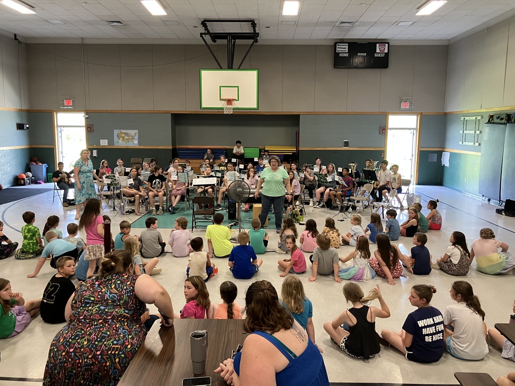 A wide, eye-level shot inside a school gymnasium where a student band is performing for an audience of younger children. The band students are seated in a large semi-circle on a teal mat under a basketball hoop, playing various woodwind and brass instruments. A female conductor in a green shirt and jeans stands at the center facing them. In the foreground, a large group of elementary school students are sitting cross-legged on the floor, watching the performance. Several adults are seated at cafeteria tables on the right side of the gym.