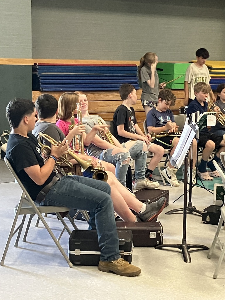 A close-up shot focusing on the brass section of a student band seated on folding chairs in a gymnasium. In the foreground, a boy wearing a black T-shirt, blue jeans, and tan boots holds a trumpet. Next to him, other students are visible holding trumpets and cornets, some chatting or adjusting their instruments. Black instrument cases are scattered on the floor beneath their chairs, and blue and green gym mats are stacked in the background.