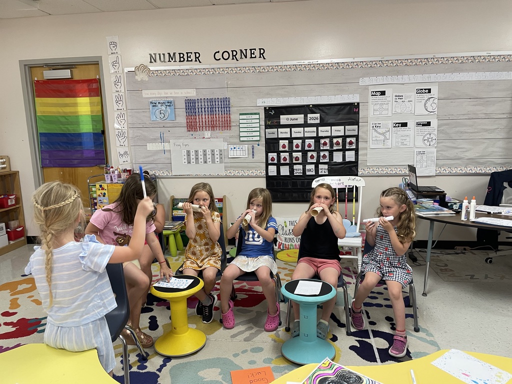 A vibrant elementary school classroom scene where a group of young girls are gathered in a circle during an activity. On the left, a girl with her blonde hair in a braided crown stands facing the group, raising a blue marker in the air as if conducting or leading. Five other girls sit in a row on chairs and colorful wobble stools; four of them are holding up rolled-up paper tubes to their mouths like kazoos or telescopes. The background features a "Number Corner" bulletin board with educational posters about maps, a calendar showing June 2026, and a rainbow-striped curtain hanging on a door. The floor is covered in a bright, multicolored patterned rug, and student desks with school supplies are visible in the foreground.