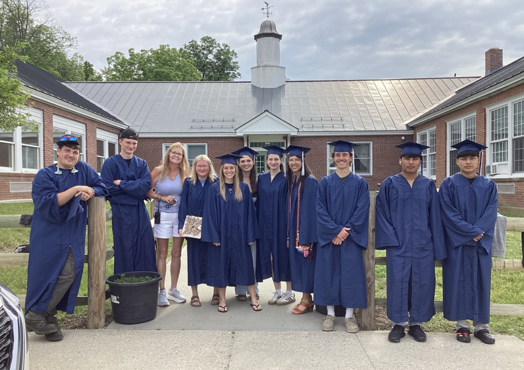 A group of Fair Haven Union High School seniors wearing blue graduation caps and gowns stand outside Orwell Village School with OVS Administrative Assistant Marcy Hart. The graduates are gathered on a walkway in front of the brick school building, smiling for a group photo as part of their return visit to their former elementary school before graduation. The sky is cloudy overhead, and the group poses in front of the school's courtyard entrance.