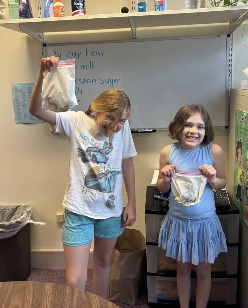 Two students proudly hold up resealable bags containing ice cream mixture. One student raises a bag overhead with an excited expression while another smiles at the camera. A whiteboard with the recipe is visible behind them.
