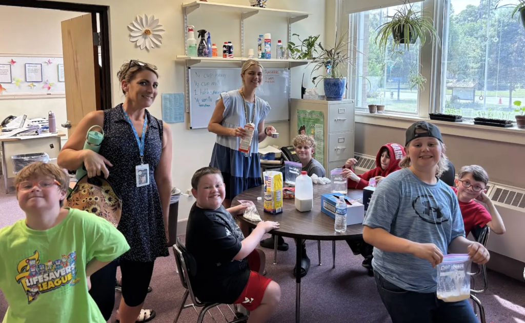 A group of students and staff pause during a homemade ice cream activity to smile for a photo. Several students hold bags containing ice cream mixture while ingredients and supplies remain set up on the table.