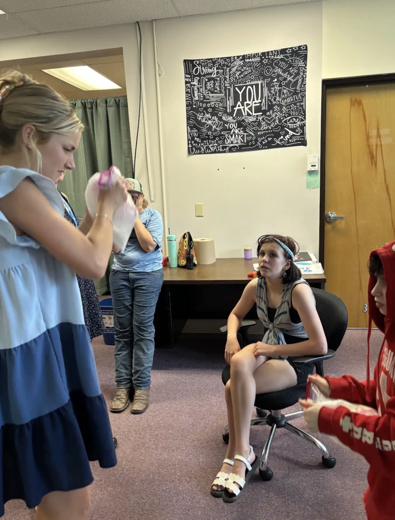 A staff member demonstrates how to shake and mix a bag of ice cream ingredients while students watch and participate. One student sits nearby observing the process as others prepare their own bags.