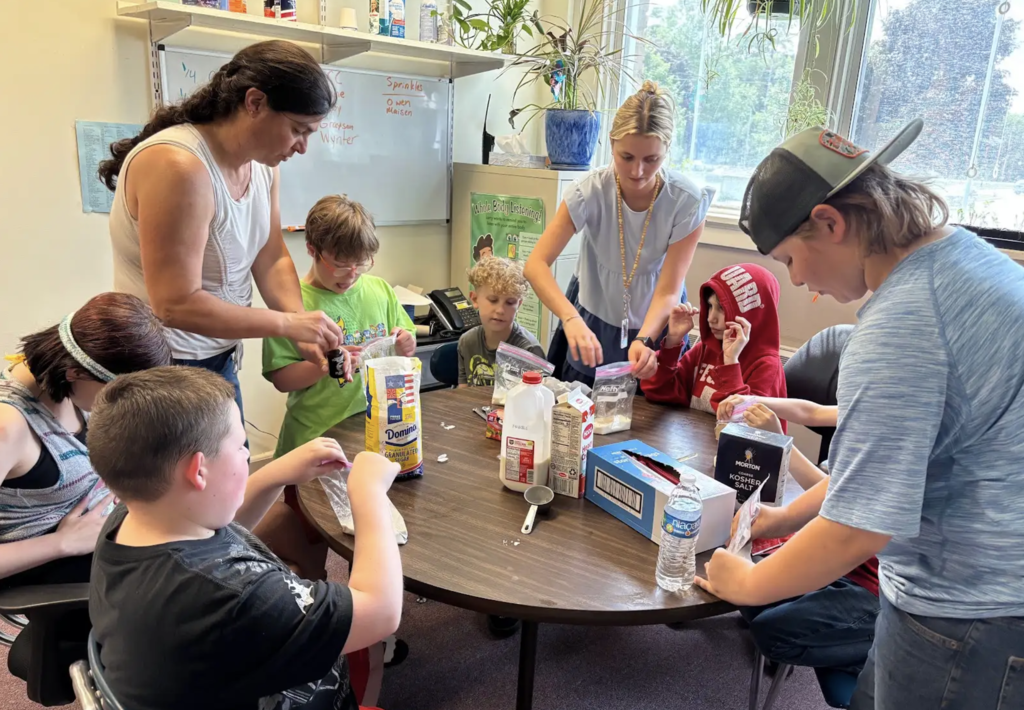 Students and staff gather around a round table preparing homemade ice cream. Ingredients including milk, sugar, salt, and measuring cups are spread across the table as participants work together to combine ingredients in plastic bags.