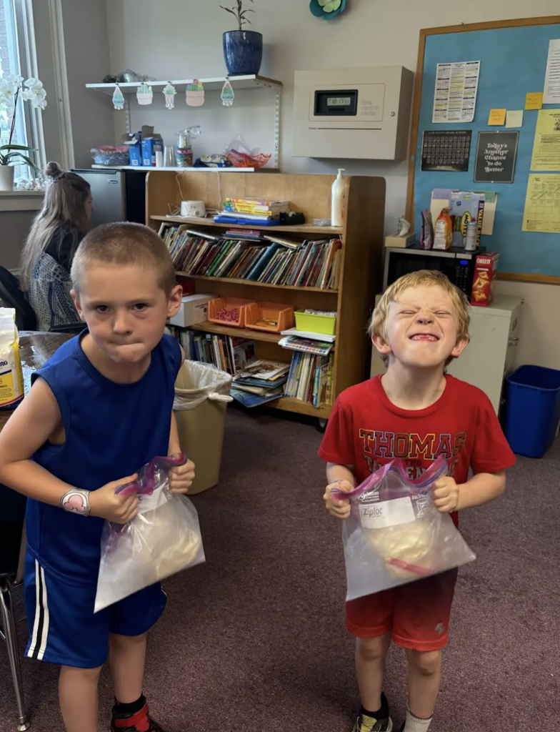 Two students stand in a classroom holding large resealable plastic bags filled with ice cream ingredients. One student in a blue sleeveless outfit looks determined, while another student in a red shirt smiles with eyes closed as they prepare to shake the bags during a homemade ice cream activity.
