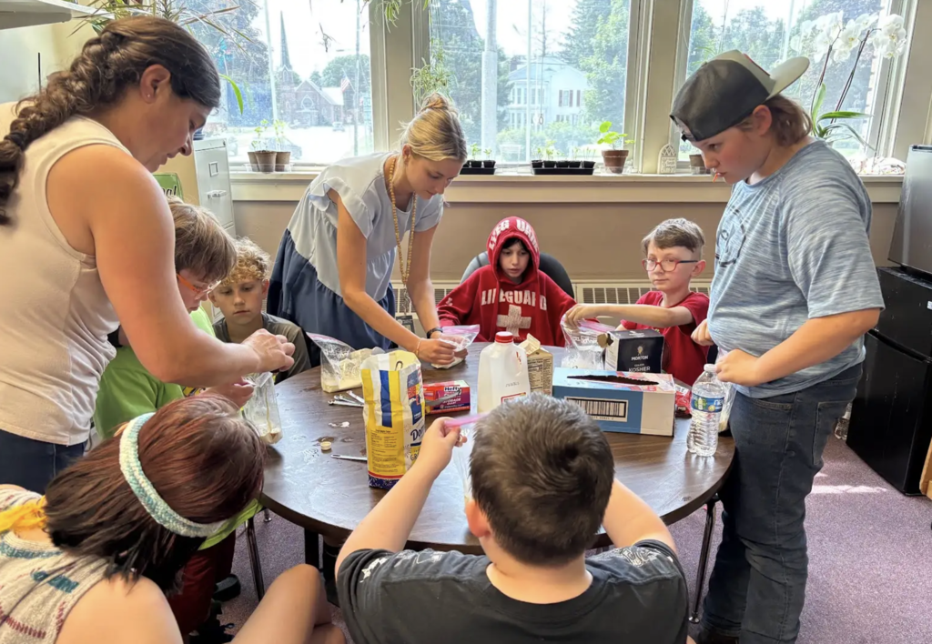 Students and staff work together around a table measuring and mixing ingredients for homemade ice cream. Containers of milk, sugar, salt, and other supplies are arranged on the table as students carefully follow directions.
