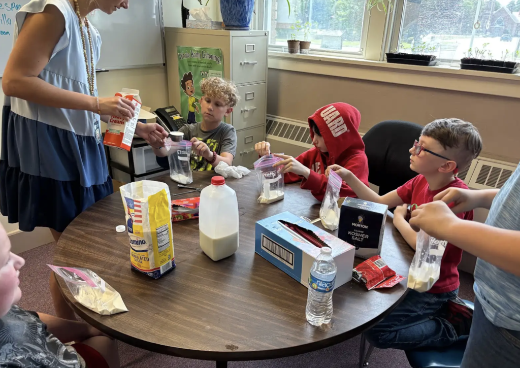 A staff member pours cream into a measuring cup while students seated around a table hold bags and watch closely. Ingredients for homemade ice cream, including sugar, milk, and salt, are visible as the group follows the recipe step by step.