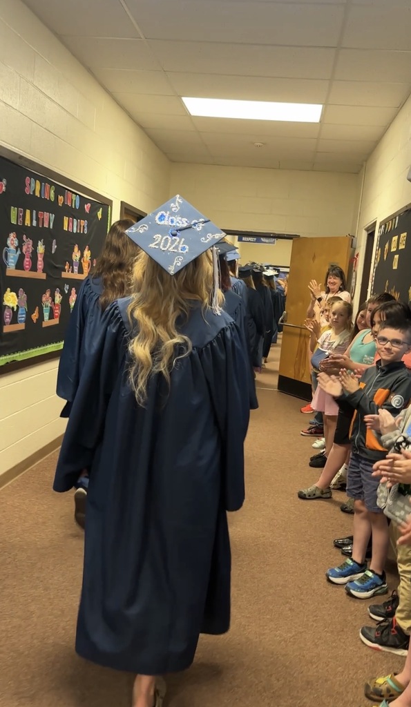 Graduating seniors walk through a school hallway while younger students and staff line the walls applauding. One senior wears a decorated graduation cap featuring “Class of 2026.”