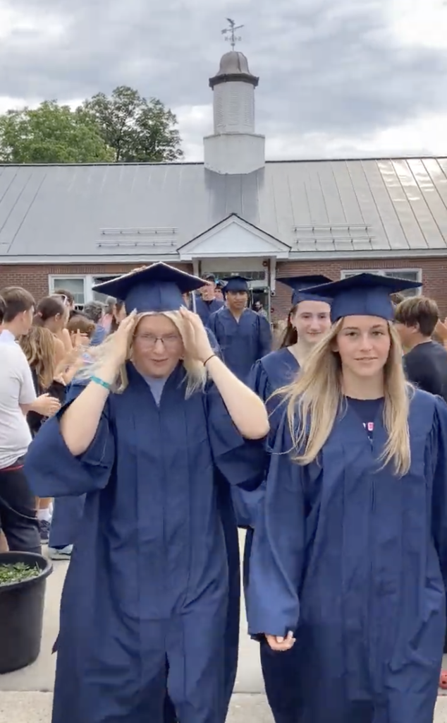 Several graduating seniors in blue caps and gowns walk through the courtyard at Orwell Village School while current students and staff line the walkway and applaud. Two seniors lead the group toward the camera.
