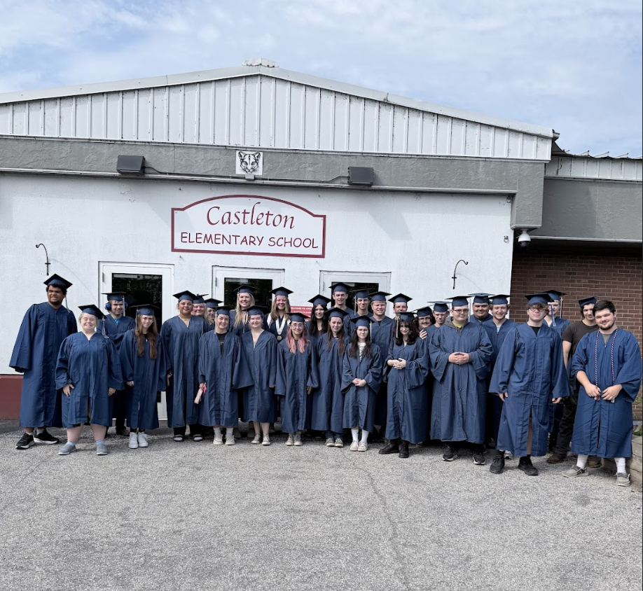 Members of the Fair Haven Union High School Class of 2026, dressed in blue graduation caps and gowns, pose for a group photo outside Castleton Elementary School. The graduates stand in front of the school entrance beneath the Castleton Elementary School sign during their senior walk visit.