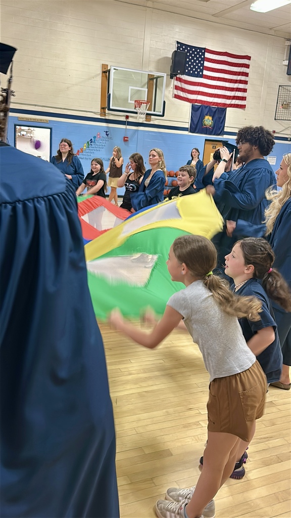 Fair Haven Union High School seniors in blue graduation caps and gowns join Castleton Elementary School students for a colorful parachute activity in the gymnasium during their senior walk visit. Elementary students and graduating seniors stand together around the parachute, lifting and lowering it while smiling and participating in the game. An American flag hangs on the wall in the background as classmates and staff look on.