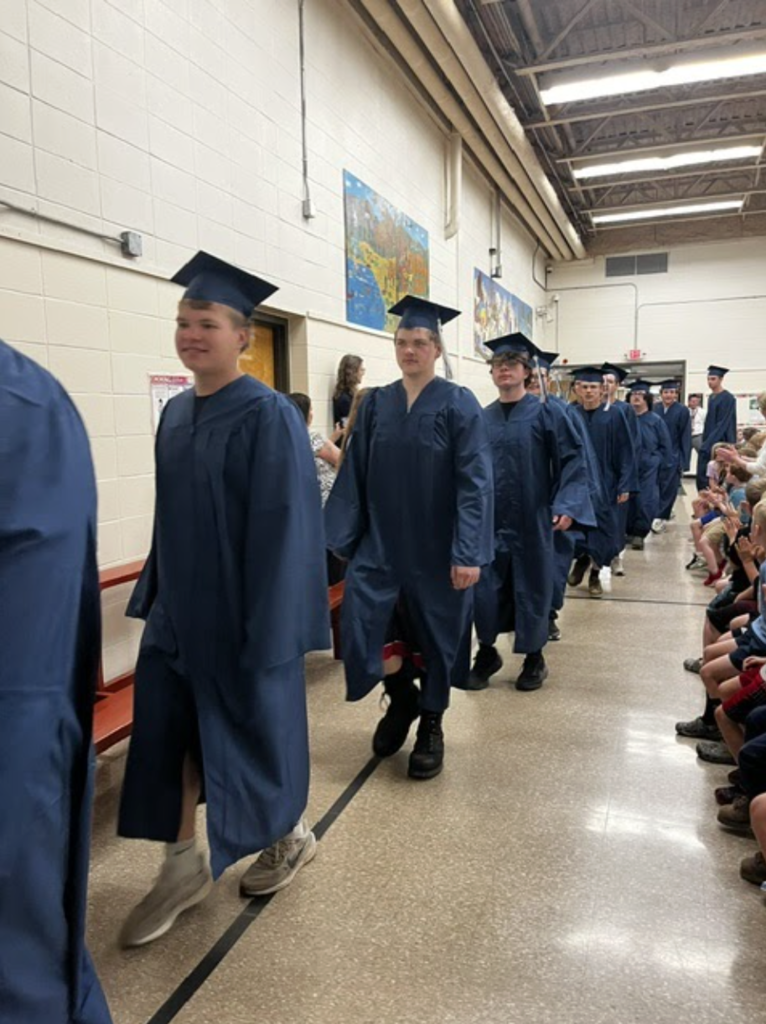 A line of Fair Haven Union High School seniors in blue caps and gowns walks through a school gymnasium. Current students sit and stand along the sides of the gym, clapping and cheering as the graduates pass by.