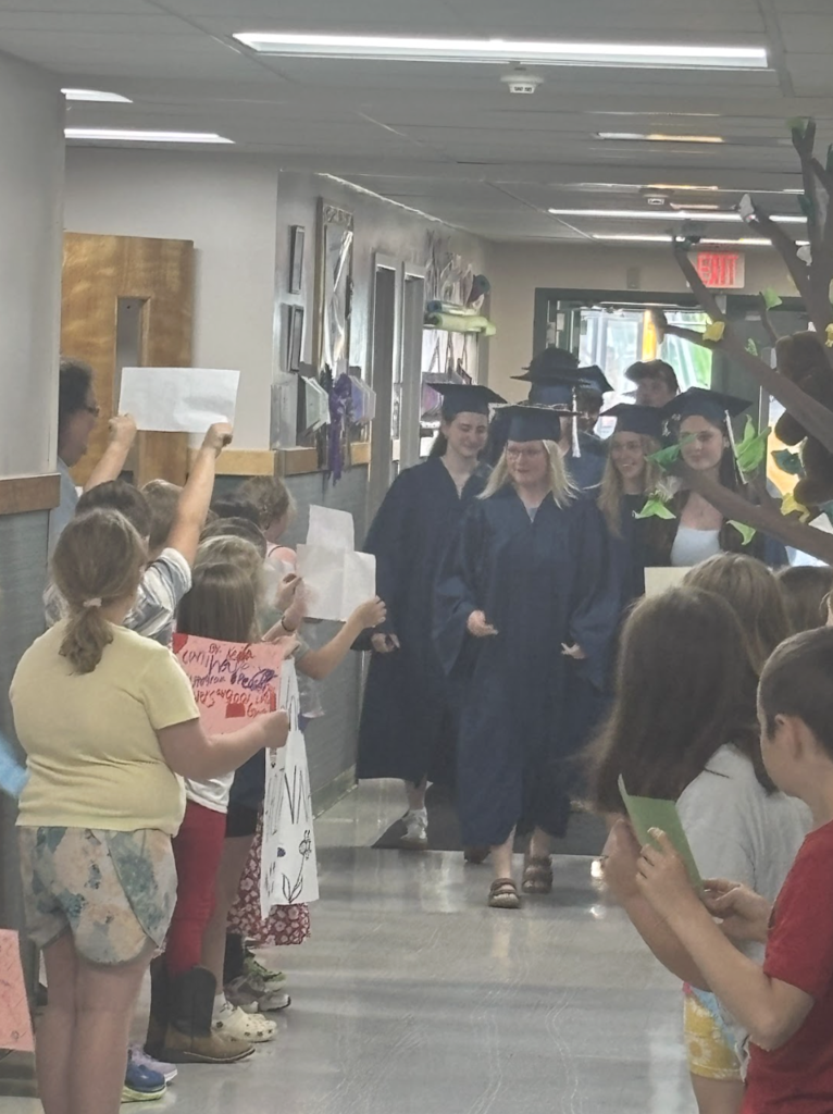 Graduating seniors in blue caps and gowns walk down an elementary school hallway lined with cheering students holding handmade signs. Younger students clap and wave as the seniors pass by.