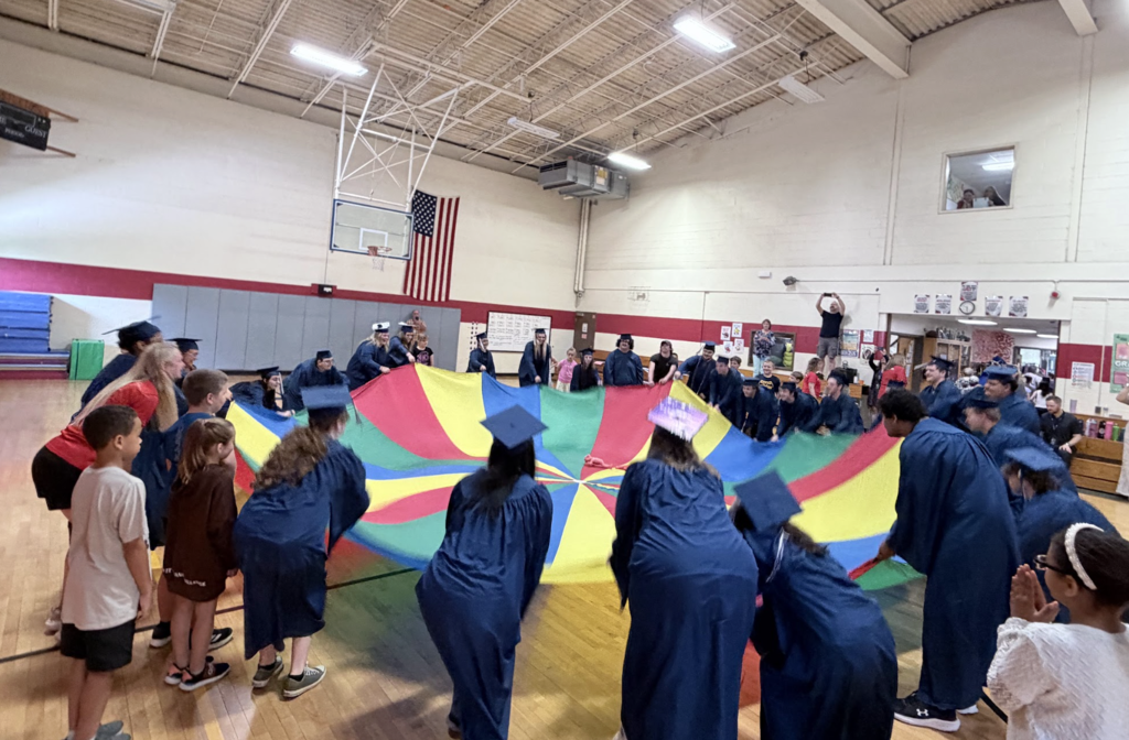 Graduating seniors in blue caps and gowns join Castleton Elementary School students in a large parachute activity in the gymnasium. Students and graduates stand around the colorful parachute together while staff and classmates watch and cheer.