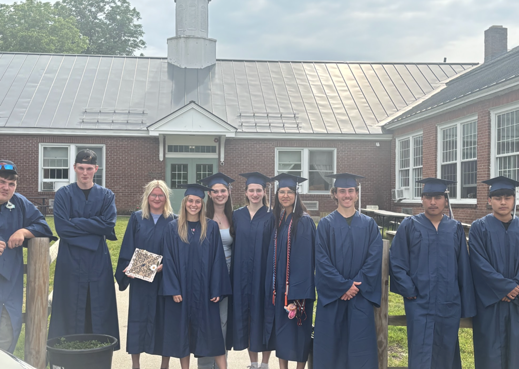 A group of Fair Haven Union High School seniors wearing blue caps and gowns pose outside Orwell Village School with a staff member. The graduates stand in front of the brick school building and smile for a photo during their senior walk visit.