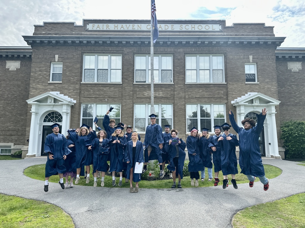 Members of the Fair Haven Union High School Class of 2026 jump into the air in front of Fair Haven Grade School. Wearing blue caps and gowns, the graduates celebrate together outside the historic brick school building.