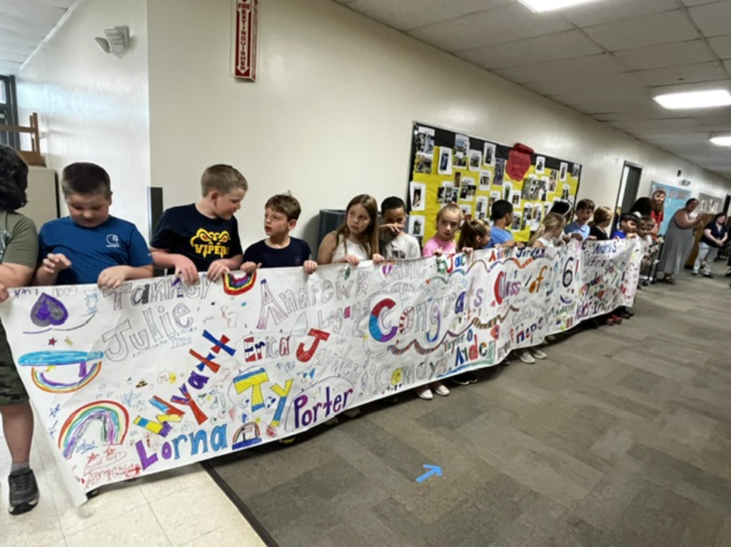 Elementary students hold a long colorful banner decorated with names, drawings, and congratulatory messages for the Class of 2026. The students stand in a school hallway waiting to greet the visiting seniors.