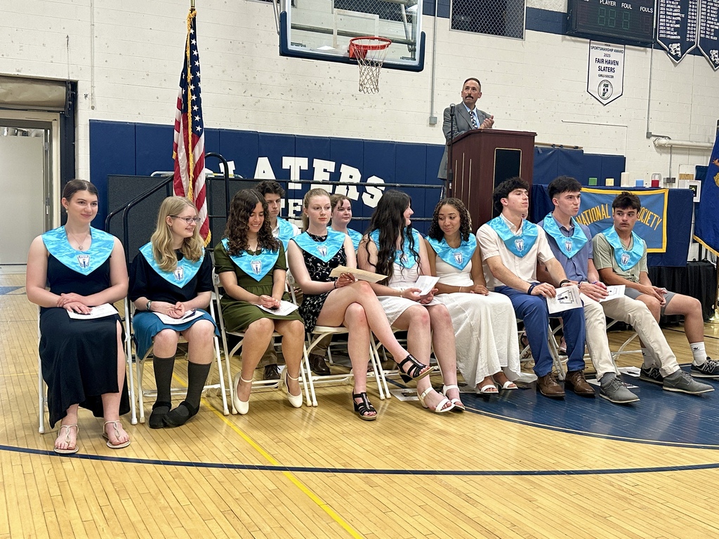 Newly inducted National Honor Society members sit together during the ceremony wearing light blue NHS stoles. Mr. Bruce stands at the podium as students listen attentively and hold their induction programs.