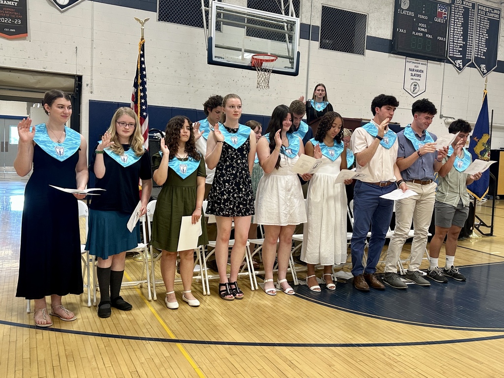 New National Honor Society inductees stand together in the gymnasium with their right hands raised as they recite the NHS pledge. Students hold ceremony programs while a current member speaks from the podium behind them.