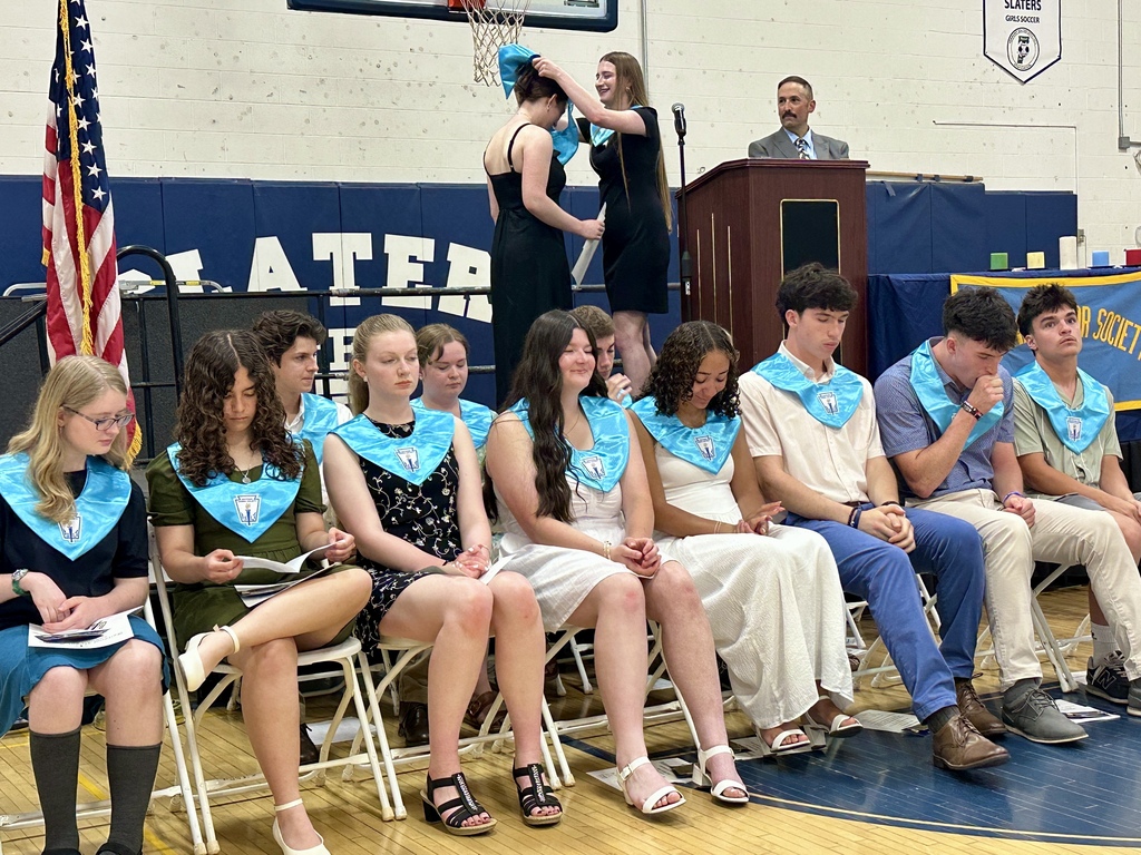 Students seated in a row during the Matthew Lyon Chapter National Honor Society Induction Ceremony wearing light blue NHS stoles. In the background, a current member places an NHS stole on a new inductee while NHS Advisor Mr. Bruce stands at a podium.