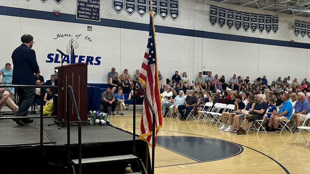 Wide view of the Fair Haven Union Middle & High School gymnasium during the Scholarship and Honor Awards Ceremony. Guests and families sit in rows of chairs while speakers and presenters stand near the stage and podium. An American flag is displayed prominently near the front of the gym.