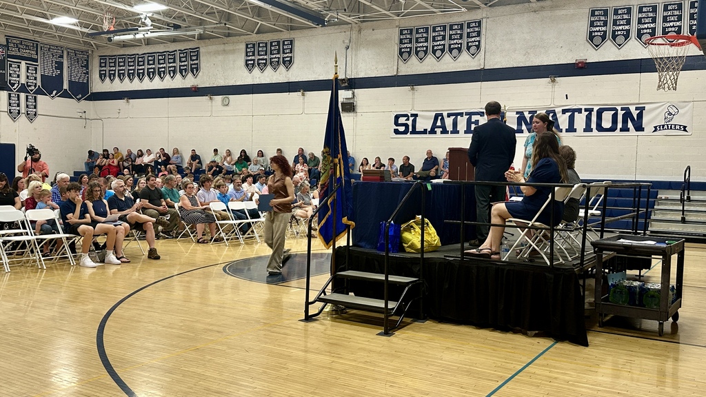 A student walks toward the stage to receive recognition during the Scholarship and Honor Awards Ceremony as family members and guests watch from their seats. Presenters and school staff stand on the stage applauding the recipient.