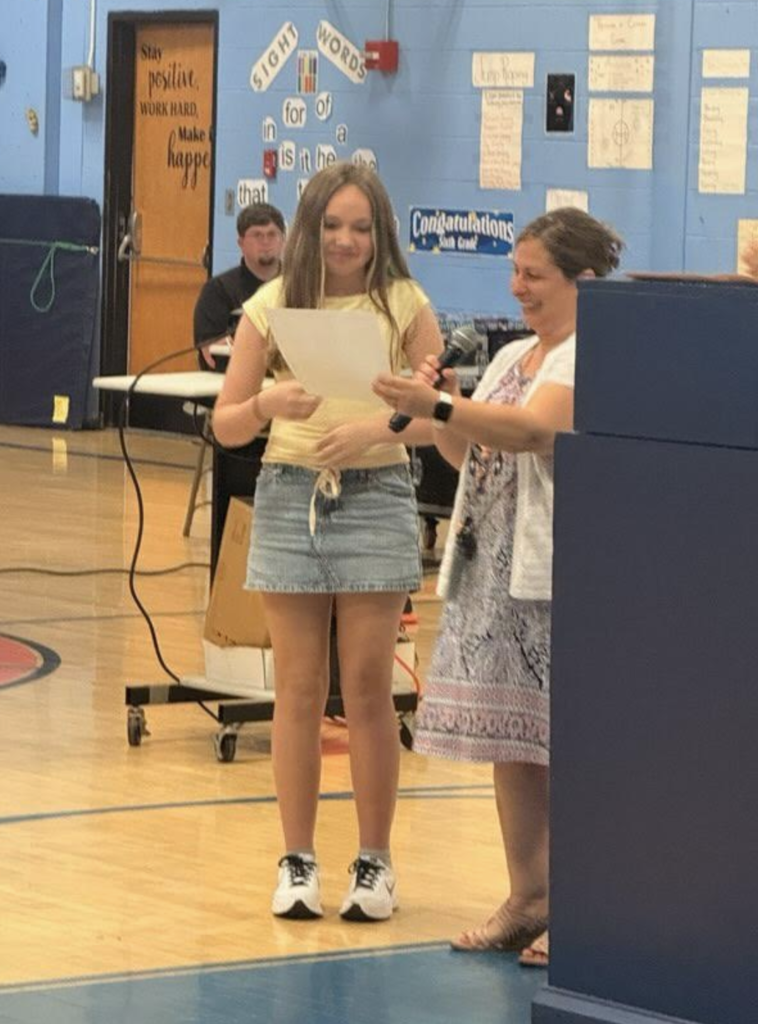 A sixth-grade student stands beside a teacher as a certificate is read aloud during the recognition ceremony in the school gymnasium.