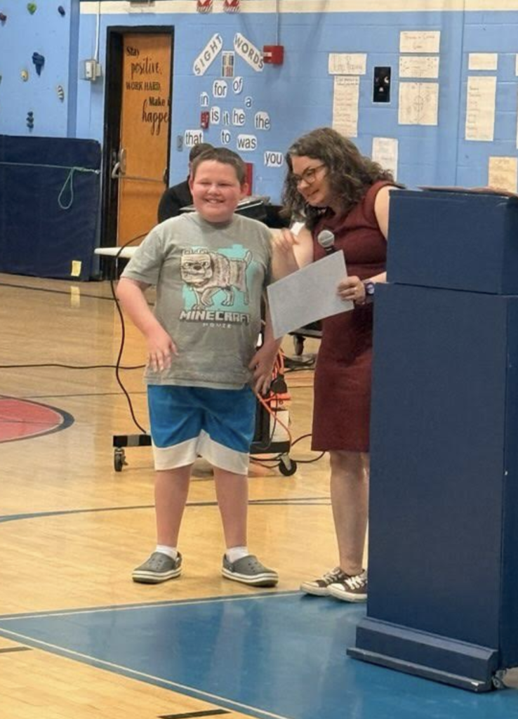A smiling sixth-grade student wearing a Minecraft-themed T-shirt stands with a teacher holding a certificate during the awards presentation.