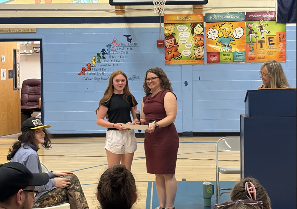 A sixth-grade student smiles while receiving a certificate during the celebration. A staff member presents the award while another staff member stands at the podium nearby.