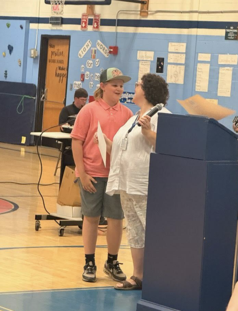A student wearing a pink polo shirt and baseball cap smiles while receiving recognition from a teacher during the sixth-grade celebration.