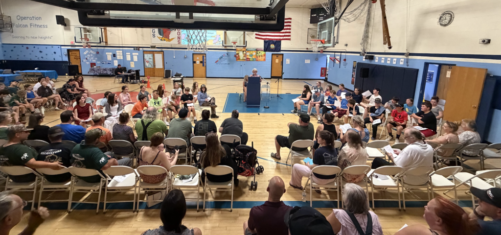 Wide view of the Fair Haven Grade School gymnasium during the 6th Grade Celebration. Students sit in a large circle while families and staff fill rows of chairs around them as a speaker stands at the podium.